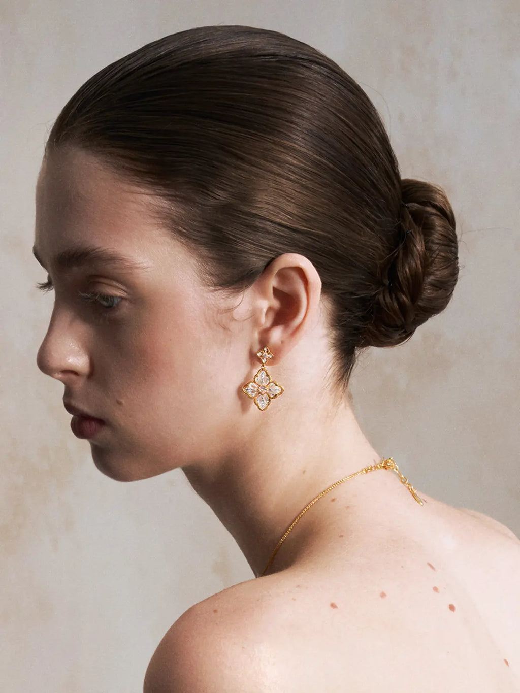 Side profile portrait of a model wearing the Tactilery Two-Tone Persimmon Calyx Earrings with hair in a low chignon, the large four-petal flower drop with matte brushed silver sand-textured petals and rope-twist gold border dangling at the lobe, paired with a delicate gold chain necklace, against a warm beige background with bare shoulders