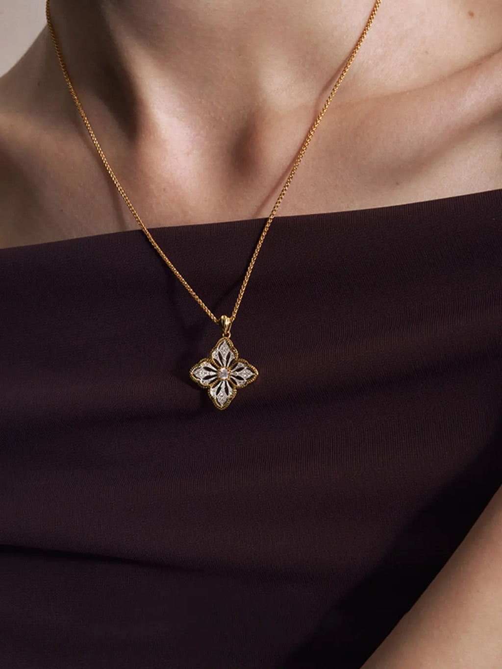 Three-quarter portrait of a model wearing the Tactilery Two-Tone Persimmon Calyx Necklace with gaze downward, the large two-tone persimmon calyx flower pendant with matte brushed silver openwork petals, fine polished gold milgrain rope border, and central prong-set large round zircon hanging at the décolletage on a polished gold twisted rope chain, paired with matching two-tone persimmon calyx bracelet and drop earrings, against a warm grey background wearing a dark draped top with dark hair slicked back